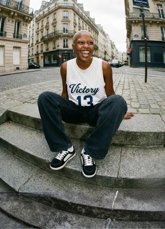 Woman wearing Barebarics Legend New York sneakers sitting on stone steps in Paris, styled with dark wide-leg jeans and a sleeveless jersey top