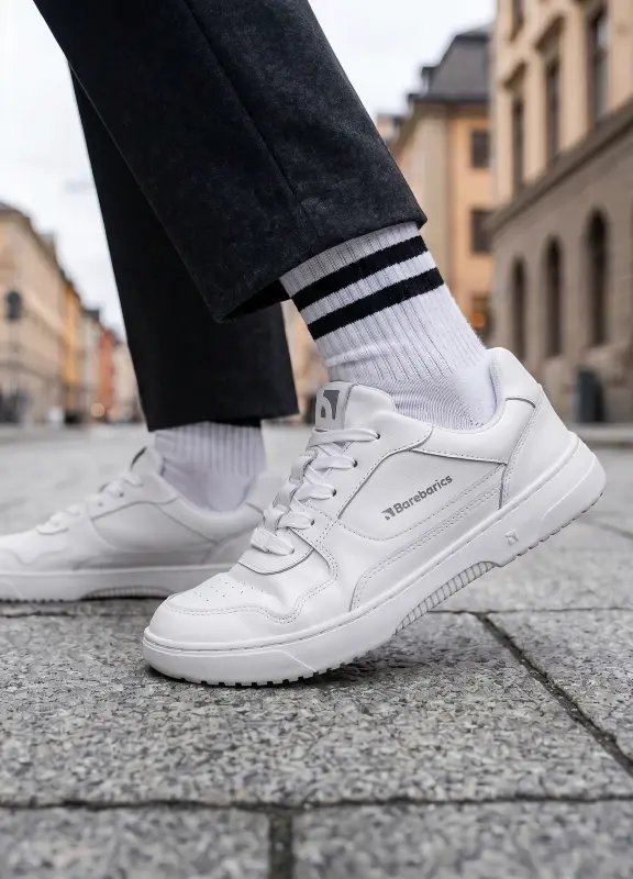 Close-up of Barebarics Zing All White sneakers on a cobblestone street, paired with striped white socks and dark pants