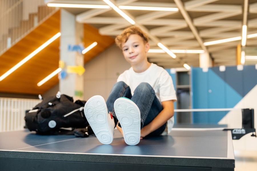 Boy sitting in a preschool hallway showing the flexible sole of white barefoot shoes for preschool