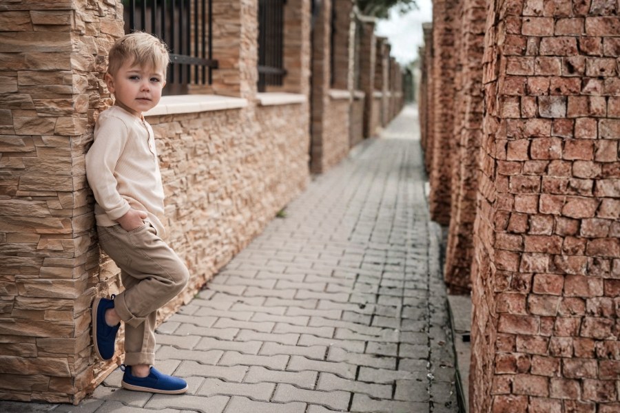 Little boy leaning against a stone wall in blue wide-toe barefoot shoes for preschool