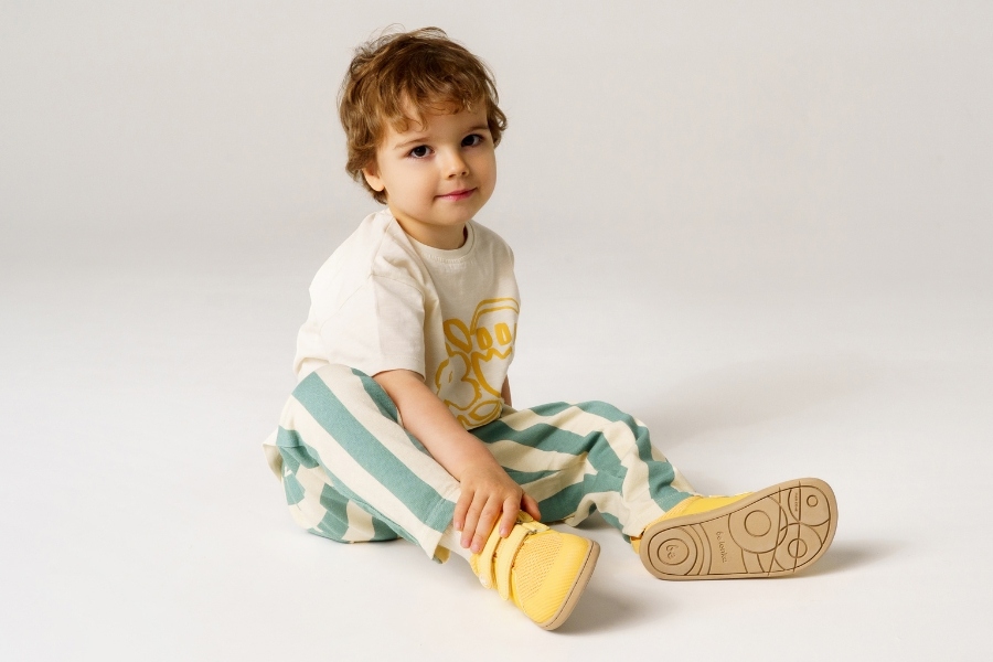 Toddler sitting and showing the flexible sole of yellow kids' barefoot shoes in size 26–34
