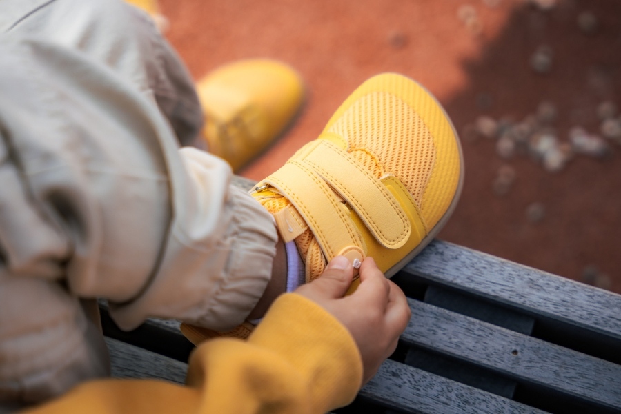 Child fastening the hook-and-loop strap on a yellow wide-toe kids' barefoot shoe, size 26–34