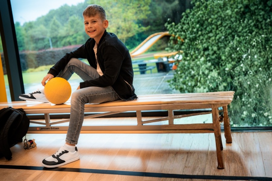 Boy with a ball sitting on a gym bench in white and black kids' barefoot shoes, size 35–40