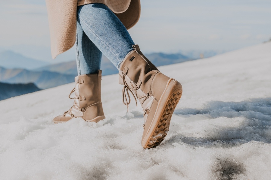 Žena v béžových dámských barefoot kozačkách kráčí po zasněženém terénu Žena v béžových dámských barefoot kozačkách kráčí po zasněženém terénu