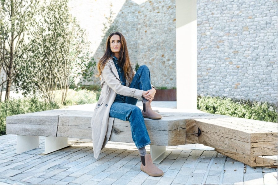 Žena v hnědých dámských barefoot kozačkách sedí na dřevěné lavičce Žena v hnědých dámských barefoot kozačkách sedí na dřevěné lavičce