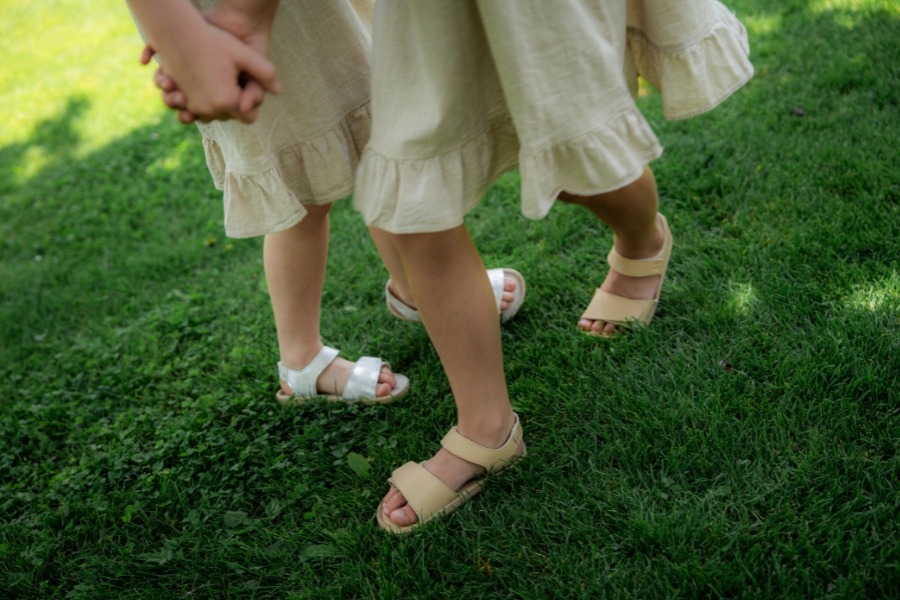 Detail nôh dvoch detí v béžových a bielych detských barefoot sandáloch na tráve