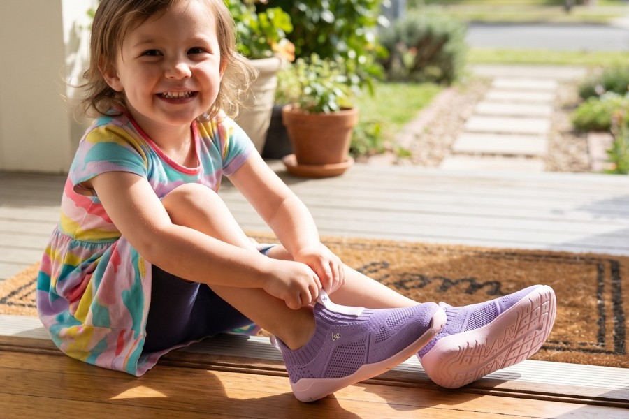 Toddler on a porch putting on purple kids' barefoot slip-ons without laces. Toddler on a porch putting on purple kids' barefoot slip-ons without laces.