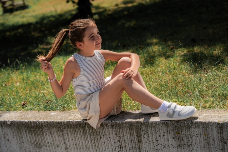 Girl sitting on a low wall in the grass in white everyday kids' barefoot shoes with hook-and-loop straps. Girl sitting on a low wall in the grass in white everyday kids' barefoot shoes with hook-and-loop straps.