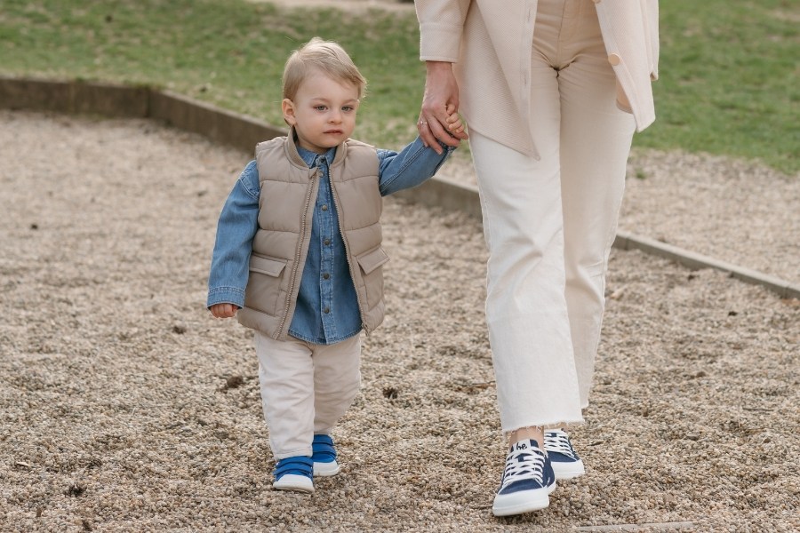 Child in blue and white kids' low barefoot shoes walking on gravel holding an adult's hand