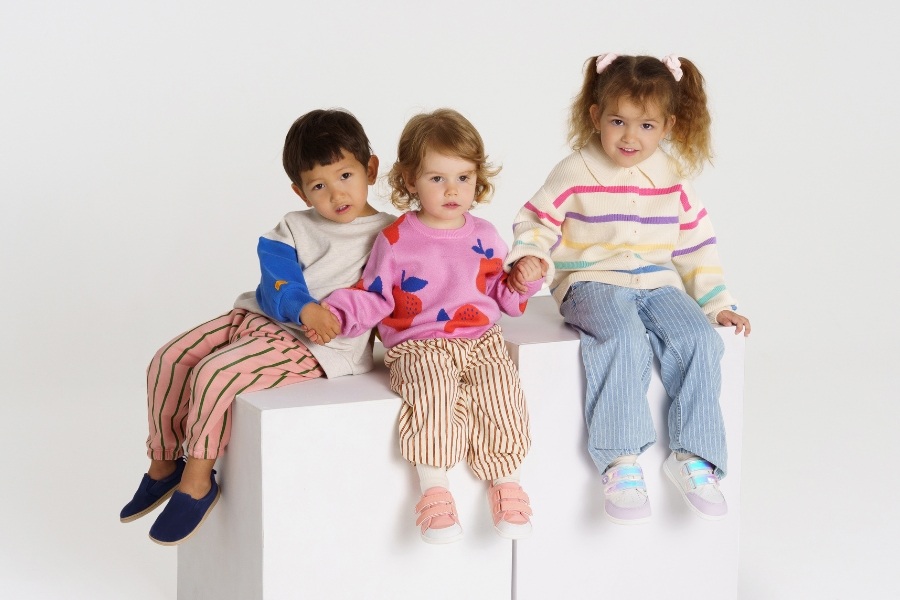 Three children in variously coloured kids' low barefoot shoes sitting on a white cube