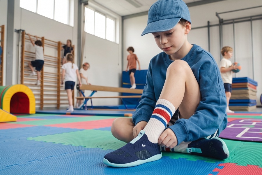 Chlapec si v telocvični obúva tmavomodré športové detské barefoot topánky s tyrkysovou podrážkou. Chlapec si v telocvični obúva tmavomodré športové detské barefoot topánky s tyrkysovou podrážkou.