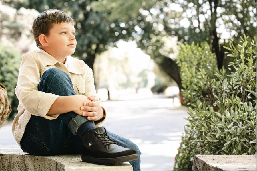 Boy sitting on a low wall in a spring park in black kids' barefoot shoes for spring.
