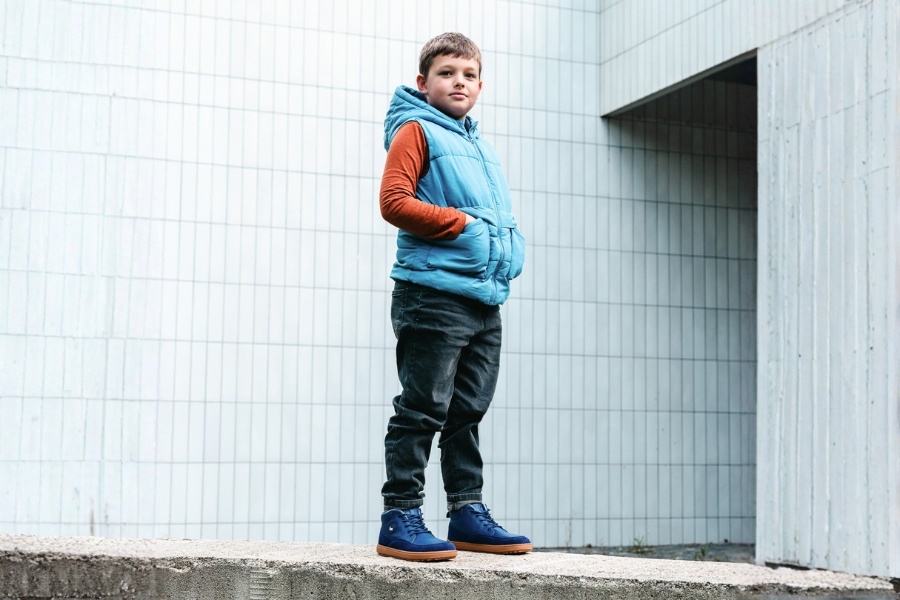Boy in a gilet standing on a wall in navy kids' barefoot shoes for spring with a gum sole.