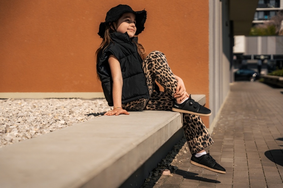 Girl in a bucket hat sitting on a concrete wall in black kids' barefoot shoes for spring.