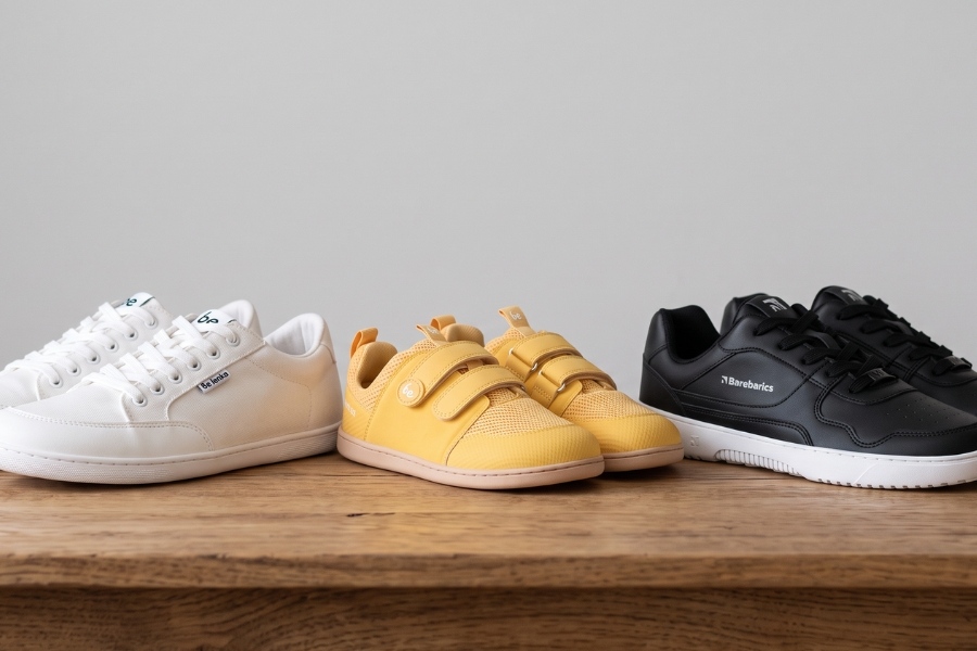 Drei Bestseller-Modelle auf einem Holzbrett: weiße und gelbe Barefoot Schuhe und schwarze Barebarics.