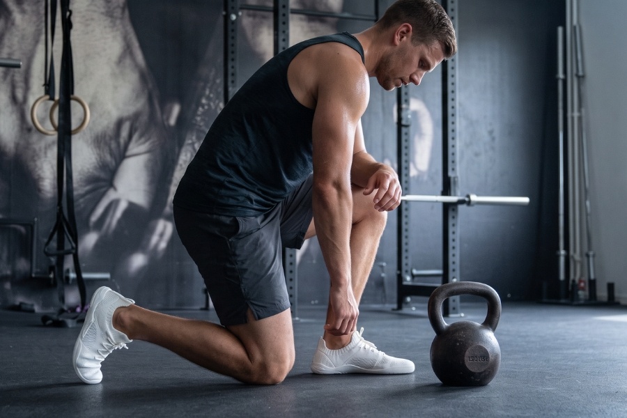 Man in white men's sports barefoot shoes kneeling by a kettlebell in a gym
