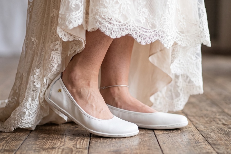 Close-up of white barefoot wedding shoes under a lace dress on a wooden floor. Close-up of white barefoot wedding shoes under a lace dress on a wooden floor.