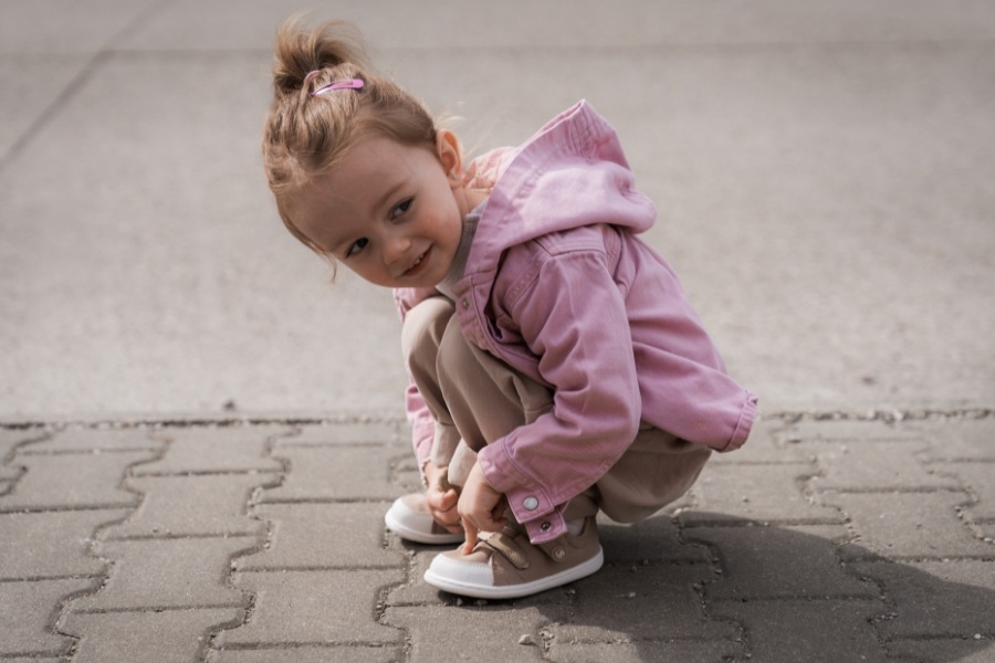 Girl in beige kids' vegan barefoot shoes fastening them on paving Girl in beige kids' vegan barefoot shoes fastening them on paving