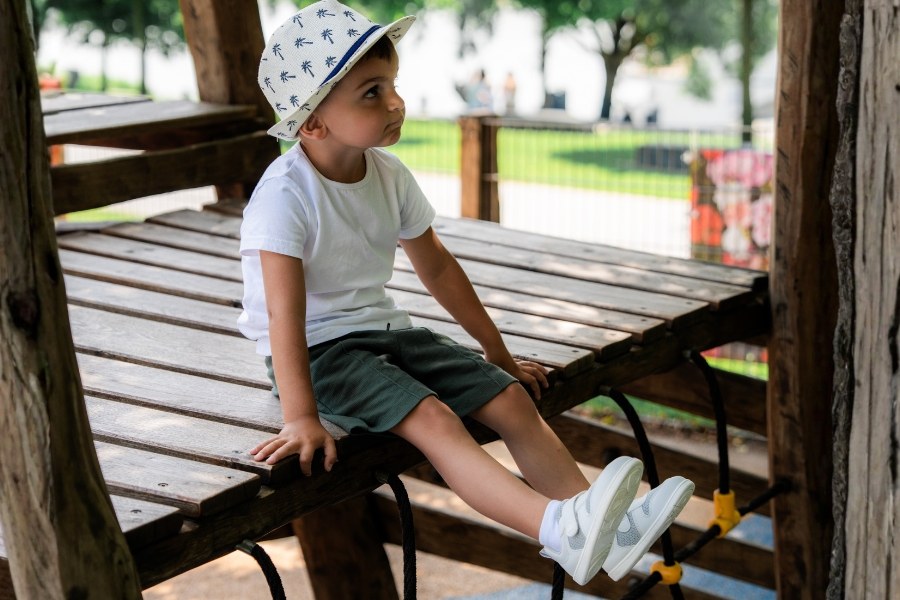 Boy in white and grey kids' vegan barefoot shoes sitting on a wooden climbing frame Boy in white and grey kids' vegan barefoot shoes sitting on a wooden climbing frame